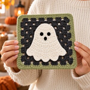 Op de afbeelding: Een vierkant gehaakt granny square met een centrale witte spookapplicatie met zwarte ogen en een zwarte mond. Het spookje is gecentreerd op een donker antracietgrijze achtergrond, omlijst door een saliegroene rand met een open steekpatroon.