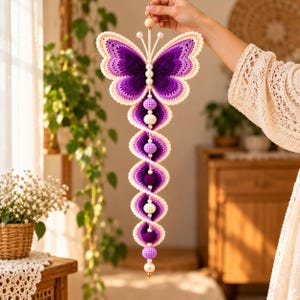May include: A hanging crochet decoration featuring a purple and cream butterfly at the top, connected to a vertical series of alternating purple and cream crocheted spheres and white beads, ending in a single large white bead at the bottom.