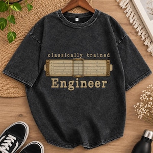 Pode incluir: Uma t-shirt cinzento-carvão com efeito de lavagem ácida, apresentando um gráfico de uma régua de cálculo vintage no centro. Acima da régua, o texto diz "classically trained" e abaixo, a palavra "Engineer" está impressa numa fonte clara.