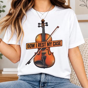 May include: A white short-sleeve t-shirt featuring a graphic of a brown violin and a bow. Across the centre of the violin is an orange rectangular sign with the black text "BOW I REST MY CASE" printed in a bold, sans-serif font style.