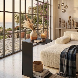 May include: A matte black metal C-shaped side table featuring a flat top surface and a lower shelf. The table holds a two-toned ceramic vase with dried pampas grass, a glass candle, and a small square coaster. A woven basket and books sit on the base.