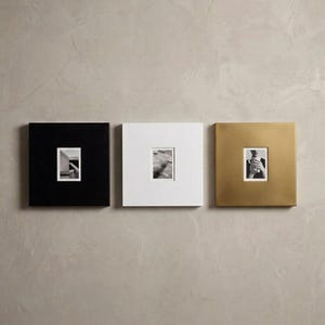 May include: Three square picture frames arranged horizontally. The left frame is matte black, the center frame is matte white, and the right frame has a metallic gold finish. Each frame features a central rectangular cutout displaying a small black and white photograph.