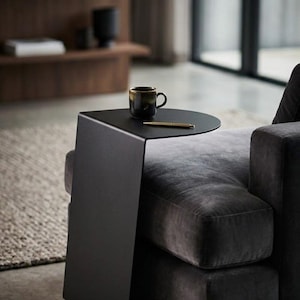 May include: A matte black metal C-shaped side table with a flat vertical base and a rounded top surface. The table holds a dark ceramic mug and a gold-toned pen, positioned directly next to a dark grey upholstered armchair with plush cushions.