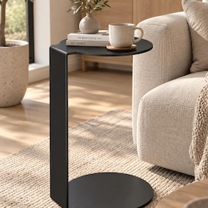 May include: A matte black C-shaped side table with a circular base and a circular top surface. The table features a continuous vertical support connecting the base to the top. On the table surface sits a beige ceramic mug on a small wooden coaster.