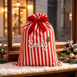Puede incluir: Una bolsa de regalo de tela con rayas verticales rojas y blancas, con un gran lazo de terciopelo rojo en la parte superior. El nombre Emily está bordado en letra cursiva blanca en el centro, junto a un pequeño icono de árbol de Navidad.