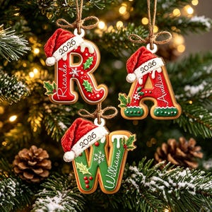 Puede incluir: Tres adornos navideños colgantes con forma de letras R, A y W, diseñados como galletas de jengibre. Cada letra presenta un acabado de glaseado rojo o verde, un gorro de Papá Noel blanco con el año 2026 y un nombre personalizado.