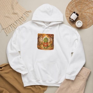 May include: A white hooded sweatshirt featuring a central graphic print of a green dinosaur wearing an orange hoodie, standing in an autumn forest with fallen leaves. The dinosaur holds a small sign with text. The hoodie has long sleeves and a front pocket.