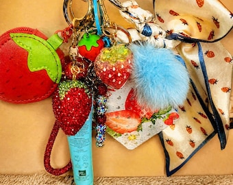 May include: A multi-charm keychain featuring a red strawberry-shaped pouch, a crystal-encrusted strawberry charm, a blue pom-pom, a small tube of lip balm, a strawberry-patterned fabric bow, and a square charm with a strawberry graphic print.