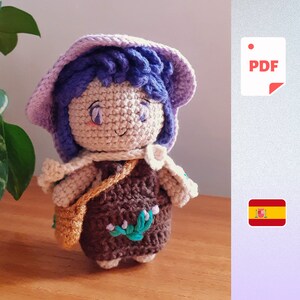 Op de afbeelding: Een gehaakte pop met paars en wit garen voor de muts en het haar. De pop draagt een bruine jurk met groene bladeren en witte bloemen. De pop houdt een klein bruin tasje vast.