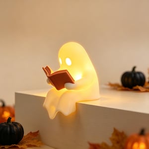 May include: A glowing, off-white, ghost-shaped lamp sitting on an edge, holding an open orange book. The lamp emits a warm, soft light from within, illuminating its rounded form and the book it holds, creating a gentle, ambient glow.