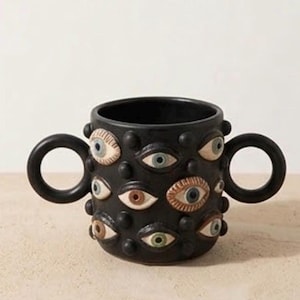 Pode incluir: Uma caneca de cerâmica preta com duas alças laterais circulares e uma superfície texturizada decorada com múltiplos motivos de olhos tridimensionais em relevo de várias cores, incluindo azul, castanho e verde, intercalados com pequenos pontos pretos.