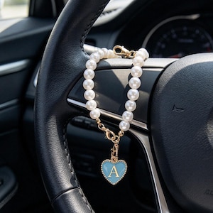 Puede incluir: Una pulsera de cuentas con perlas blancas esféricas unidas por un cierre de metal dorado. Un dije en forma de corazón con un centro de esmalte azul claro y una letra A dorada está sujeto a la pulsera mediante un pequeño clip de metal.