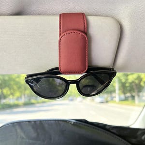 Könnte beinhalten: Ein matter, terrakottafarbener Brillenhalter-Clip aus Kunstleder, der an einer hellgrauen Auto-Sonnenblende befestigt ist. Der Clip verfügt über einen Magnetverschluss und hält eine schwarze Brille mit dunklen Gläsern sicher fest.