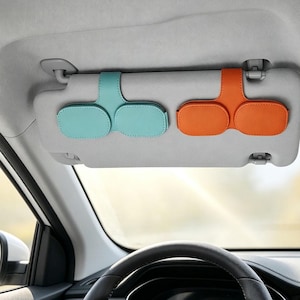 Puede incluir: Dos soportes para gafas de sol para el parasol del coche fijados a un parasol gris. Un soporte es de color azul claro y el otro es de color naranja. Ambos soportes tienen un diseño en forma de T con dos compartimentos ovalados para gafas.