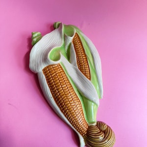May include: A ceramic wall hanging shaped like two ears of corn with yellow kernels, light green husks, and a brown silk tassel at the base. The piece features a glossy finish and realistic textures, depicting the corn partially wrapped in leaves.