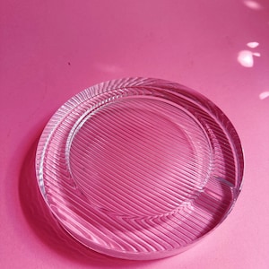 May include: A circular clear glass coaster featuring a textured surface with fine, parallel diagonal ridges. The glass is transparent, allowing light to pass through and create subtle shadows, with a smooth, raised outer rim and a recessed centre.