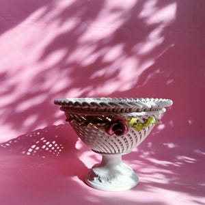 May include: A white ceramic pedestal bowl featuring an intricate woven lattice design around the body and a braided rim. The bowl is decorated with a small, sculpted pink rose and green leaves attached to the side, resting on a flared base.