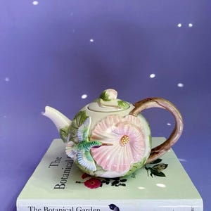 May include: A ceramic teapot featuring a three-dimensional pink flower and a green hummingbird on its side. The teapot has a light green body, a brown handle, and a lid with a small sculpted bud. It rests on a book titled The Botanical Garden.