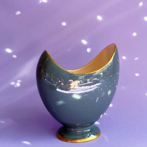 May include: A dark teal ceramic vase with a glossy finish and a curved, asymmetrical rim. The vase features a pedestal base and a cream-coloured interior. Both the top rim and the base are accented with a thin, metallic gold-coloured trim.