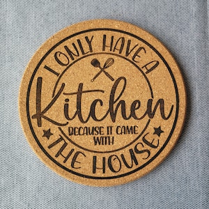 Può includere: Un sottopentola rotondo in sughero con il testo "Ho solo una cucina perché è arrivata con la casa" e un paio di utensili da cucina incrociati.