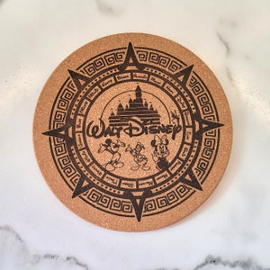 May include: A round cork coaster with a black and white design featuring the Walt Disney World logo, Cinderella's Castle, and various Disney characters. The coaster has a decorative border with a repeating Greek key pattern.