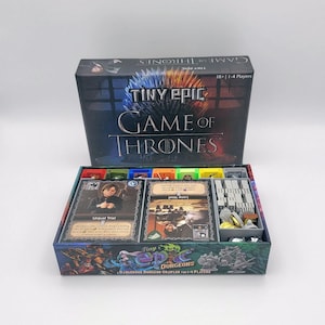 Puede incluir: Una caja gris y negra con un inserto de juego para los juegos de mesa Tiny Epic Dungeons y Tiny Epic Game of Thrones. La caja tiene un esquema de color verde, azul y rojo. La caja está abierta y el inserto de juego es visible. El inserto tiene compartimentos para cartas, fichas y dados. La caja está etiquetada como "Tiny Epic Dungeons" y "Tiny Epic Game of Thrones".