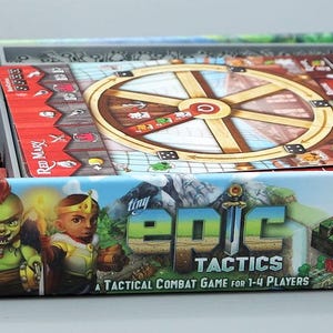 Może przedstawiać: Pudełko gry planszowej "tiny Epic Tactics" z elementami gry, kostkami i planszą. Pudełko przedstawia postacie fantasy, a plansza ma okrągły kształt. Gra jest dla 1-4 graczy.