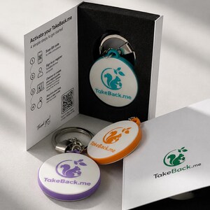 Op de afbeelding: Drie ronde TakeBack.me-sleutelhangers met eekhoornlogo's in turquoise, oranje en paars, gepresenteerd in een wit doosje met zwarte voering. De binnenkant van de doos bevat gedrukte activeringsinstructies, inclusief een QR-code.
