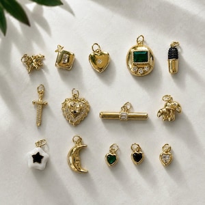 May include: A collection of gold-toned metal charms featuring various shapes including a dinosaur, a sword, a crescent moon, a mushroom, a toggle bar, and several hearts with green, black, and clear stones, each equipped with a small jump ring.