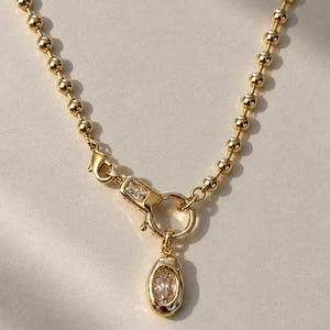Könnte beinhalten: Eine goldfarbene Kugelkette mit einem zentralen Verschlussmechanismus, der mit einem kleinen rechteckigen, klaren Stein verziert ist. Ein am Verschluss befestigter runder Ring hält einen ovalen Anhänger mit einem facettierten, klaren Stein.
