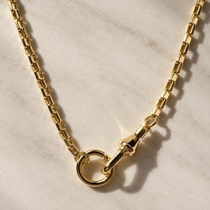May include: A gold-toned box chain necklace featuring a polished finish. The chain is connected to a circular ring clasp, which is secured by a matching gold-toned swivel lobster claw mechanism, creating a continuous and uniform metallic link design.