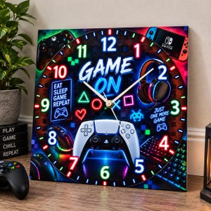 Peut inclure: Une horloge murale carrée au thème de jeu vidéo vibrant avec des graphismes style néon, incluant une manette, un casque et le texte "GAME ON", "EAT SLEEP GAME REPEAT" et "JUST ONE MORE GAME" avec des aiguilles blanches lumineuses.