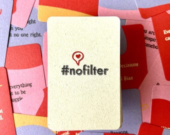 Puede incluir: Primer plano de una tarjeta con el texto "#nofilter" en negro y un corazón rojo con un alfiler en la parte superior. La tarjeta es beige y tiene una superficie texturizada. La tarjeta está rodeada de otras tarjetas con varios textos y colores.