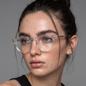 May include: Clear, transparent, round-framed spectacles with a subtle gold-toned metal accent at the hinges. The frames feature a polished, translucent finish that highlights the bridge and temples, providing a clean, neutral look for the wearer.