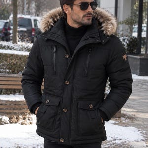 May include: A black hooded parka featuring a faux fur trim, a high collar, and a front zipper closure with additional buttons. The jacket includes two vertical chest pockets with zippers and two lower flap pockets with button closures.
