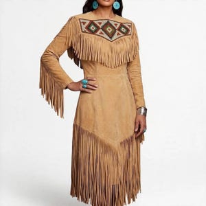 May include: A tan suede dress featuring long sleeves with fringe detailing, a round neckline, and a geometric patterned panel across the chest. The dress includes a V-shaped fringe overlay on the bodice and a long fringe hemline at the bottom.