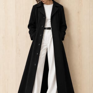 May include: A long black coat with a collared neckline and a front button closure, worn over a white top and white trousers with a black belt. The coat features long sleeves and a straight, floor-length hemline, creating a structured silhouette.