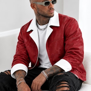 Könnte beinhalten: Eine rote Jacke mit weißem Kragen und weißen Manschetten, getragen über einem schlichten weißen T-Shirt. Die Jacke hat einen Reißverschluss und horizontale Nahtdetails. Dazu werden eine schwarze Jeans mit Rissen und Silberschmuck getragen.