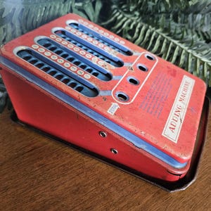 May include: A vintage red metal adding machine with blue sliding number tracks and circular input holes. The device features the text ADDING MACHINE printed on the top surface, along with a small arrow pointing to the word TOTAL near the controls.