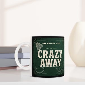 May include: A dark green ceramic mug with a white handle featuring the text "ONE MATCHA A DAY KEEPS THE CRAZY AWAY" in a bold, vintage-style font. The design includes a line-art illustration of a matcha whisk and a small bowl on the front.