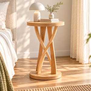 May include: A light-toned wooden side table featuring a circular top and a base composed of three curved, intersecting legs that meet at a solid round floor plate. The table surface holds a white dome-shaped lamp and a small vase with greenery.