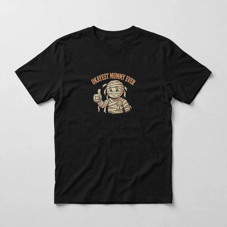 May include: A black short-sleeved t-shirt featuring a graphic of a cartoon mummy character giving a thumbs up. The mummy is wrapped in bandages and wears a small award ribbon. Above the character, the text reads OKAYEST MUMMY EVER in orange.
