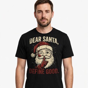 May include: A black short-sleeved t-shirt featuring a graphic of Santa Claus wearing a red hat and winking while holding a finger to his lips. The text above reads DEAR SANTA, and the text below reads DEFINE GOOD in a distressed red font.
