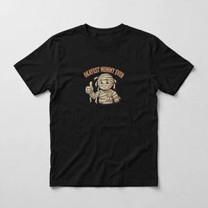 May include: A black short-sleeved t-shirt featuring a graphic of a cartoon mummy character giving a thumbs up. The mummy is wrapped in bandages and wears a small award ribbon. Above the character, the text reads OKAYEST MUMMY EVER in orange.