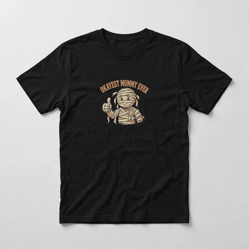 May include: A black short-sleeved t-shirt featuring a graphic of a cartoon mummy character giving a thumbs up. The mummy is wrapped in bandages and wears a small award ribbon. Above the character, the text reads OKAYEST MUMMY EVER in orange.