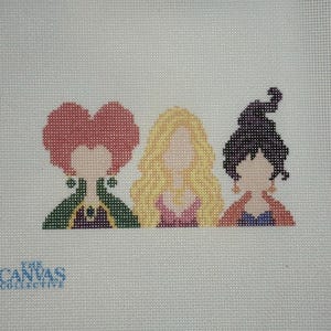 May include: A needlepoint canvas featuring three stylized figures with no facial features. The left figure has red hair and a green cloak, the center figure has long blonde hair and a pink top, and the right figure has dark hair and an orange shawl.