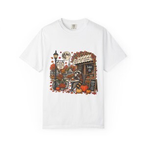 Może przedstawiać: Biały t-shirt z kolorową grafiką przedstawiającą szopa pracza siedzącego przy stoliku przed sklepem o nazwie Luke's. Scena zawiera latarnię uliczną, rower, dynie i jesienne liście. Tekst na grafice brzmi Stars Hollow, Coffee Pie Friends oraz Luke's Coffee Pies & Good Vibes.