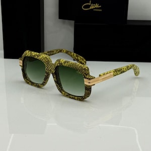 Puede incluir: Gafas de sol Cazal Legends con montura cuadrada y estampado de piel de serpiente en amarillo y negro. Las gafas tienen lentes verdes degradadas y detalles metálicos dorados en las varillas. El nombre Cazal Legends aparece en el estuche negro.