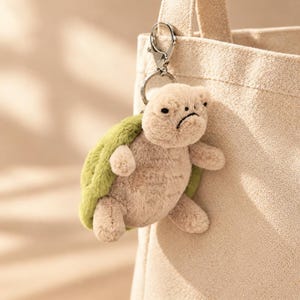 May include: A plush turtle keychain featuring a soft beige body and a textured green shell. The turtle has a small, embroidered face with a neutral expression and is attached to a silver-toned metal lobster clasp and ring for easy attachment.