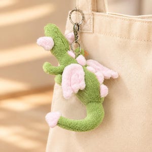 Può includere: Un portachiavi a forma di drago in peluche verde con ali rosa morbide, dettagli rosa sulle orecchie e una coda con punta rosa. La creatura imbottita è fissata a un moschettone in metallo argentato e a un anello circolare, appesa a una borsa in tela beige.